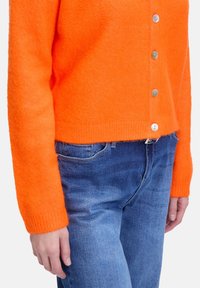 Oranžový vlněný cardigan s ribbeným lemem a dlouhými rukávy, zdobený stříbrnými knoflíky vpředu, v kombinaci s modrými džínovými kalhotami.