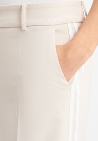 Beige broek met een gladde textuur, voorzien van een zijzak en witte gestreepte accenten langs de naad. Geborduurd logo boven de zak.