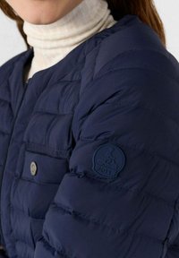 Giacca imbottita blu navy con design trapuntato, dotata di chiusura con zip, tasca con bottone a pressione e toppa a trama sulla manica con logo.