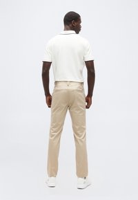 Άνδρας που στέκεται γυρισμένος πλάτη, φοράει λευκό πόλο  με κοντά μανίκια, μπεζ παντελόνι και λευκά αθλητικά παπούτσια σε απλό φόντο.