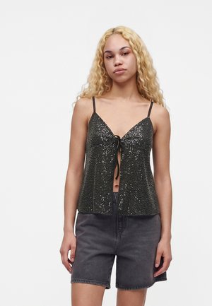 Jeune femme aux longs cheveux blonds porte un top sans manches noir à sequins avec un nœud à l'avant et un short en jean gris foncé.