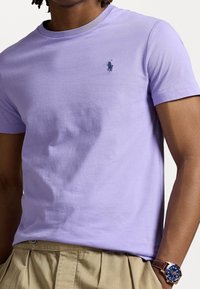 Man som bär en ljuslila kortärmad t-shirt med en liten mörkblå polo-spelare logotyp, beige plisserade byxor och en blå urtavla armbandsklocka med brunt läderarmband.