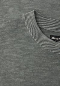 Gros plan sur un pull gris à col rond côtelé montrant une texture de tricot fine et une étiquette de vêtement noire partiellement visible.