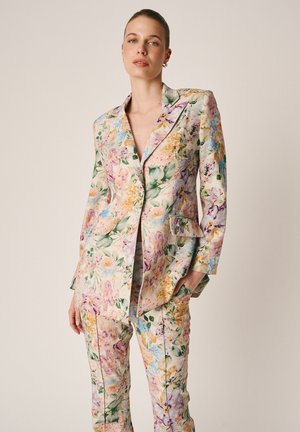 Mujer vestida con un blazer de estampado floral en tonos pastel y pantalones a juego, posando con una mano en el bolsillo sobre un fondo liso.