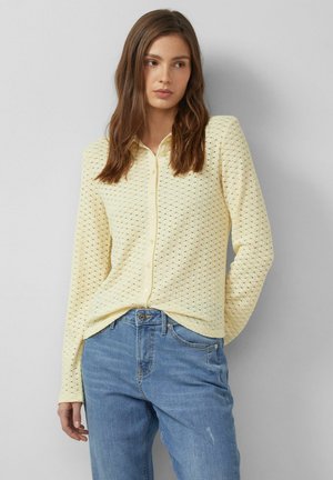 Kobieta o długich brązowych włosach, ubraną w bladożółty, fakturowany sweter zapinany na guziki oraz jasnoniebieskie dżinsy, stojąca na tle jednolitej szarej ściany.