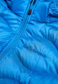 Blå quiltet jakke med sort lynlås og "Peak Performance" broderet logo på stoffet.