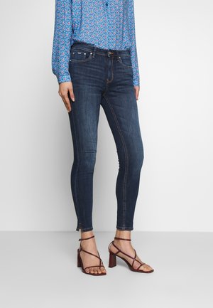 Jean skinny bleu foncé au design ajusté, ornés de coutures discrètes et d'un léger zip au niveau des chevilles, associés à des talons avec lanières.