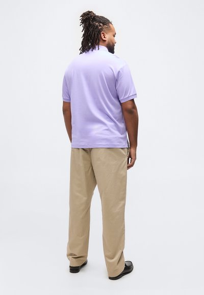 Άνδρας με ντραντλοκς, με την πλάτη γυρισμένη, φοράει ανοιχτό μωβ πόλο πουκάμισο, μπεζ παντελόνι και μαύρα επίσημα παπούτσια σε απλό λευκό φόντο.