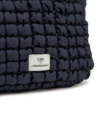 Sac texturé bleu marine avec un design matelassé, une étiquette blanche proéminente avec du texte noir et une finition lisse.