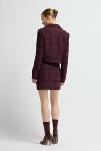 Completo in tweed bordeaux con blazer corto e gonna aderente. Presenta un motivo a quadri, polsini a coste, abbinato a tacchi alti e calzini.
