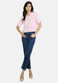 Röksleeve rozā teksturēta blūze ar apkakli, kurai ir pieci zelta krāsas pogas, kopā ar augsto vidukļa zilu džinsu biksēm.