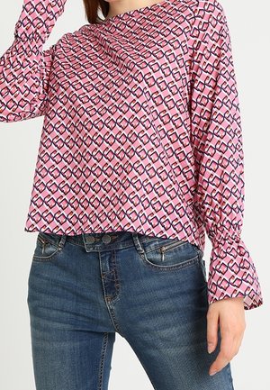 Femme portant un chemisier à manches longues rose à motif géométrique et un jean bleu foncé, posant avec un bras levé.