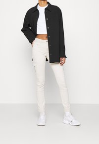 Czarna koszula z guzikami noszona na białej bluzce crop top, w połączeniu z beżowymi spodniami cargo z bocznymi kieszeniami oraz białymi butami sportowymi.