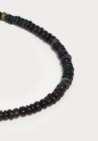 Κοντινό πλάνο από μια καμπυλωτή αλυσίδα από σκούρες, στρογγυλές χάντρες όπαλ με λεπτές αναλαμπές πράσινου, κόκκινου και κίτρινου σε λευκό φόντο.