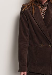Blazer in velluto a coste marrone con ampi revers, bottoni doppi e una sottile texture. Indossato su un top a fantasia con stampa leopardata.