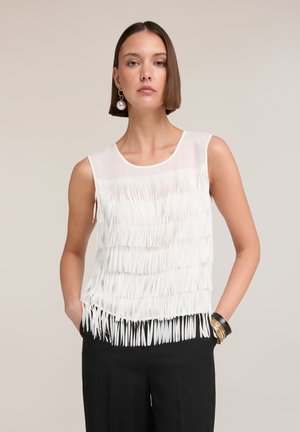 Młoda kobieta o krótkich brązowych włosach nosi bezrękawnik z białymi frędzlami, czarne spodnie, dużą perłową kolczyk oraz nałożone bransoletki, pozując z rękami w kieszeniach.