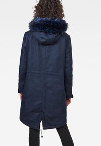 Marinblå kappa med huva och fodrad konstpäls, med en texturerad yta, lång stil och dragsko vid nederkant. Bakvy visas.