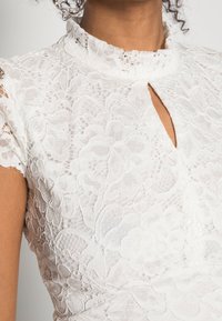 Robe en dentelle blanche ornée de motifs floraux, avec un col haut et un détail en trou de serrure au niveau du décolleté ; texture transparente avec des manches courtes.