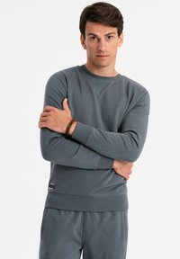 Grauer Baumwoll-Sweatshirt mit langen Ärmeln, gerippten Bündchen und rundem Halsauschnitt. Hat eine dezente Textur und ein kleines Markenlogo am Saum.