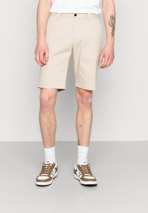 Beige Baumwollshorts mit geradem Schnitt, umgeschlagenem Saum und einem einzelnen braunen Knopfverschluss. Kombiniert mit weißen, gerippten Socken und braunen Sneakers.