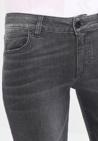 Jeans de mezclilla gris con una textura suave, que presentan un diseño clásico de cinco bolsillos, costuras visibles y un cierre de botón único en la cintura.