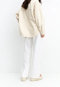 Beige oversized shirt met een ronde zoom, gecombineerd met witte wijde broek en beige platte sandalen. Een gladde textuur van de stof is zichtbaar.