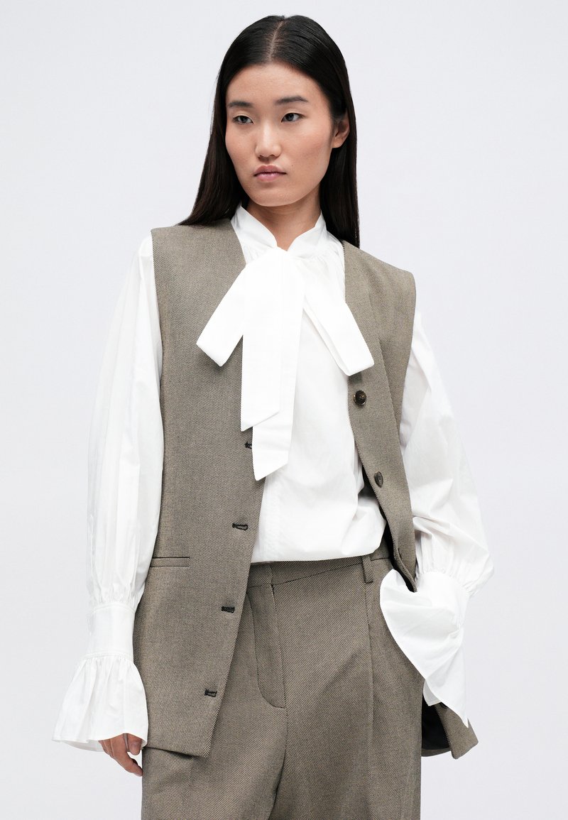Lichtbruine vest met een gestructureerd ontwerp, voorzien van een voorclosuresluiting met knopen en zakken, gelaagd over een witte blouse met strik en gestructureerde broek.
