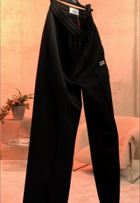 Mustad kõrge vöökohaga püksid riputatud riidepuule, vöökohal tikitud detail, taustal potitaim ja beež tugitool.