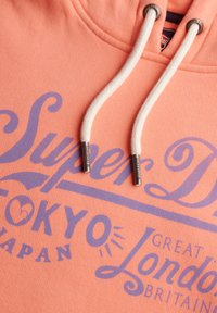 Różowa bluza z kapturem z fioletowym nadrukiem graficznym z napisem "Super D" oraz "Tokio, Japonia." Sznurki w kolorze białym z metalowymi końcówkami. Miękka tekstura.