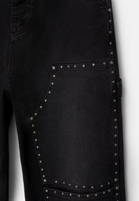 Pantalon en jean noir avec une grande poche latérale bordée de clous métalliques argentés disposés uniformément autour des bords de la poche.