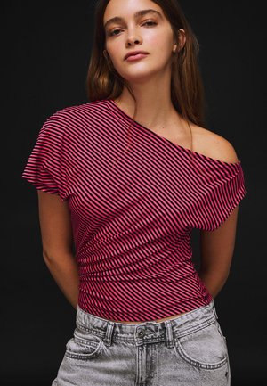 Mlada žena u ružičastoj i crnoj prugastoj majici s otkrivenim ramenima i svijetlo sivim trapericama, stoji ispred crne pozadine.