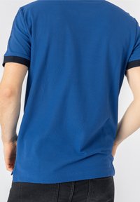 Muž v modrém polotričku s černým lemem na rukávech a tmavých džínách, zobrazený zezadu na jednoduchém bílém pozadí.