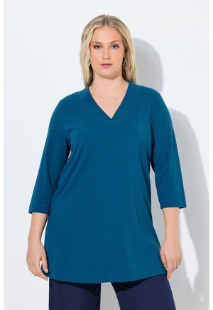 Blonde Frau, die eine türkisfarbene Tunika mit V-Ausschnitt, dreiviertellangen Ärmeln und marineblaue Hose trägt, vor einem schlichten weißen Hintergrund stehend.