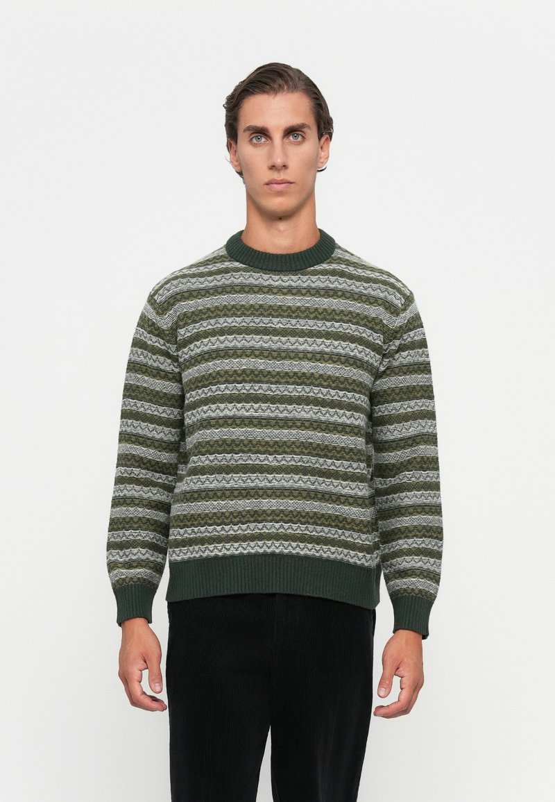 Młody mężczyzna stoi przodem w zielono-szarym, paskowanym swetrze dzianinowym i czarnych spodniach na białym tle.