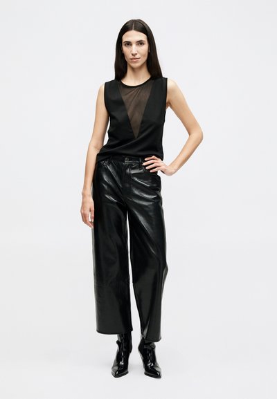 Woman with long dark hair stands wearing sleeveless black top with sheer V-panel, wide-leg black leather pants, and black pointed boots.