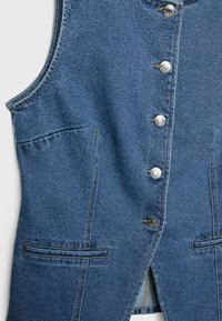 Denimli yelek, orta mavi renkte, gümüş düğmeli, iki ön cepli ve kenarları yırtmaçlı düz bir kesime sahip.