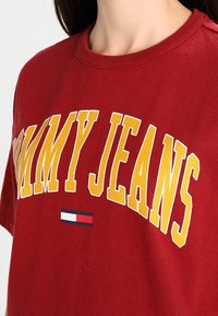 Camiseta de algodón roja con gran texto amarillos "TOMMY JEANS" y un pequeño acento de logo azul, blanco y rojo. Diseño de manga corta y cuello redondo.