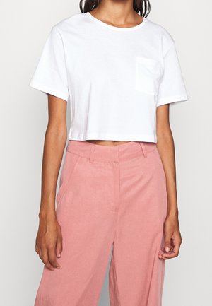 Frau trägt ein weißes, kurzärmeliges Crop-T-Shirt mit Brusttasche und hoch taillierte, lockere rosa Hose vor einfachem Hintergrund.