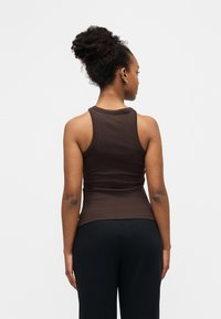 Femme aux cheveux bouclés attachés en chignon, portant un débardeur sans manches marron foncé et un pantalon noir, debout de dos devant un fond blanc uni.