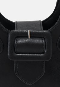 Gros plan d'une boucle de sac en cuir noir avec une sangle cousue et une épingle argentée maintenant la sangle dans le cadre rectangulaire de la boucle.