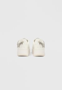 Λευκά δερμάτινα sneakers με πλατφόρμα, εμφανιζόμενα από πίσω, με ασημένιες αλυσίδες ως διακοσμητικά και διακριτικό μοτίβο μονόγραμμου στο πίσω μέρος.