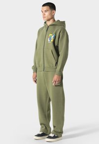 Oliven grøn zip-up hoodie med frontlommer og farverig grafik på brystet; parret med matchende sweatpants og sorte sneakers.