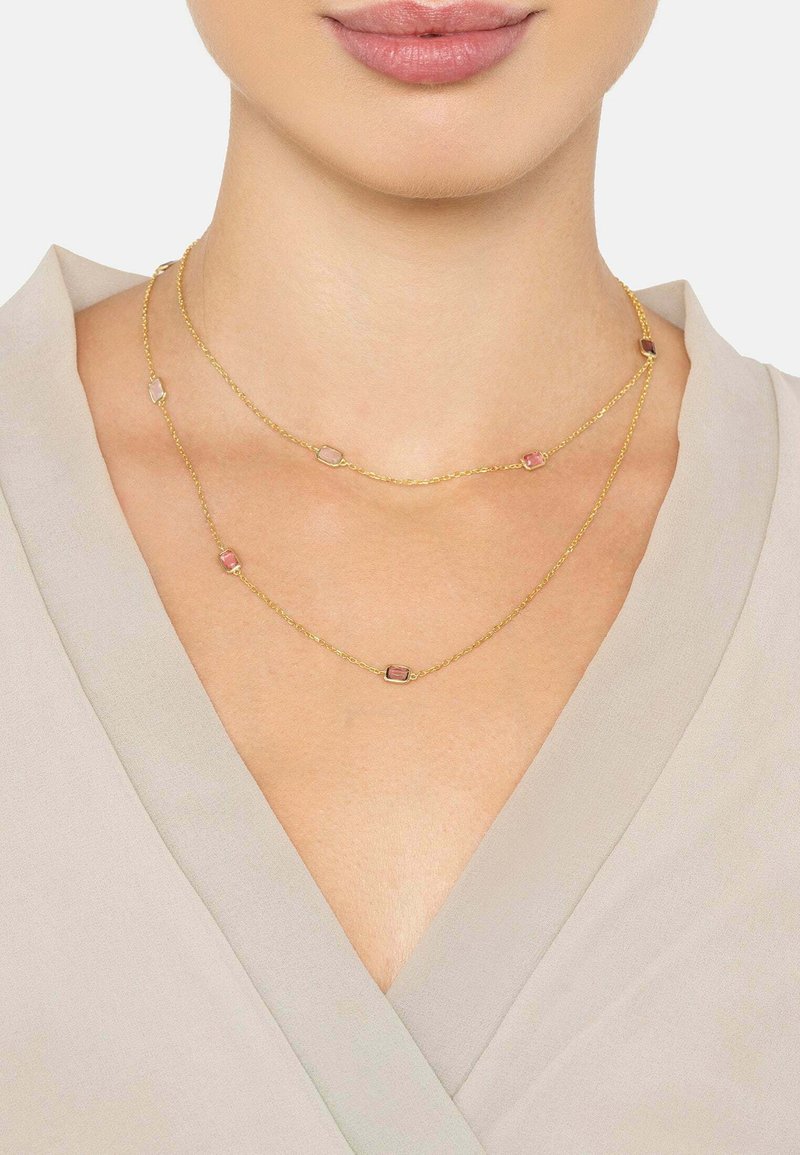 Gold-tone layered necklace featuring rectangular pink and clear stones, set on fine chain. Worn against a light-colored v-neck top.
