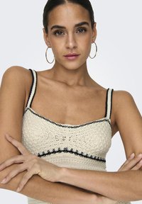 Crochet knit top in cream with black accents. Features spaghetti straps and a textured design with a fitted silhouette.
