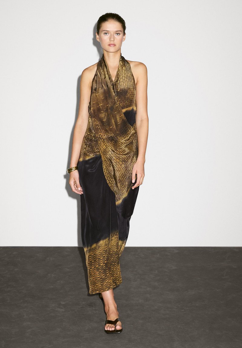 Robe portefeuille marron et noire avec un motif texturé, fabriquée en tissu léger. Présente un col halter et un design fluide, portée avec des sandales.