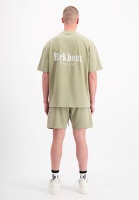 Olijfgroen katoenen t-shirt met wit logo op de achterkant, gecombineerd met bijpassende shorts. Model draagt witte sneakers en sokken met zwarte accenten.