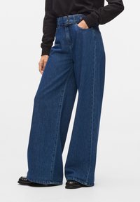 Jean en denim à jambes larges dans un bleu foncé. Pr�ésente une taille haute, des passants de ceinture et une finition texturée subtile. Tissu lisse avec une coupe décontractée.