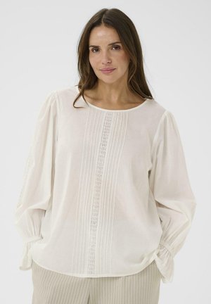 Kobieta w białej bluzce z długim rękawem ozdobionej koronką i plisowaniami, w połączeniu z beżowymi spodniami w prążki, stojąca na jednolitym tle.