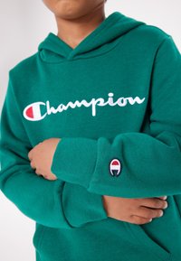 Felpa verde realizzata in tessuto morbido, con il logo bianco "Champion". Include una patch con il logo sulla manica con accenti rossi e blu.