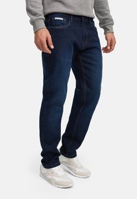 Mann iført mørkeblå jeans, grå genser og hvite og beige joggesko, stående mot en ensfarget hvit bakgrunn.